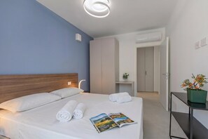 A hotel room with a bed, a bedside table with a magazine, a potted plant, and a wardrobe. - Nua Boutique Apartaments 2 Towncentre, Finale Ligure, Italy (Finale Ligure)