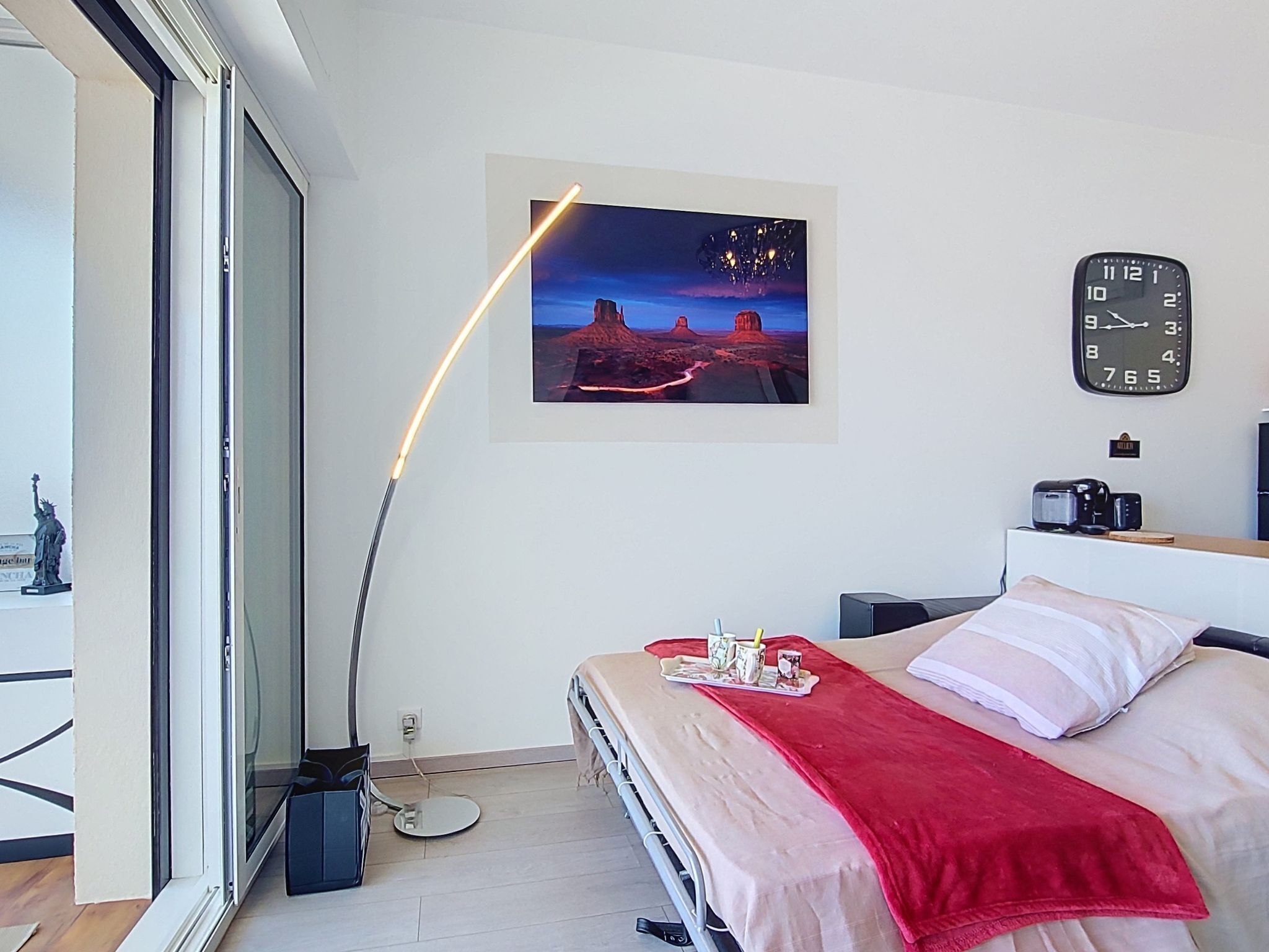 Een slaapkamer met een bed, een rode deken, een klok, een foto aan de muur en een raam met uitzicht.