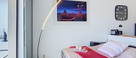 A bedroom with a bed, a red blanket, a clock, a picture on the wall, and a window with a view.