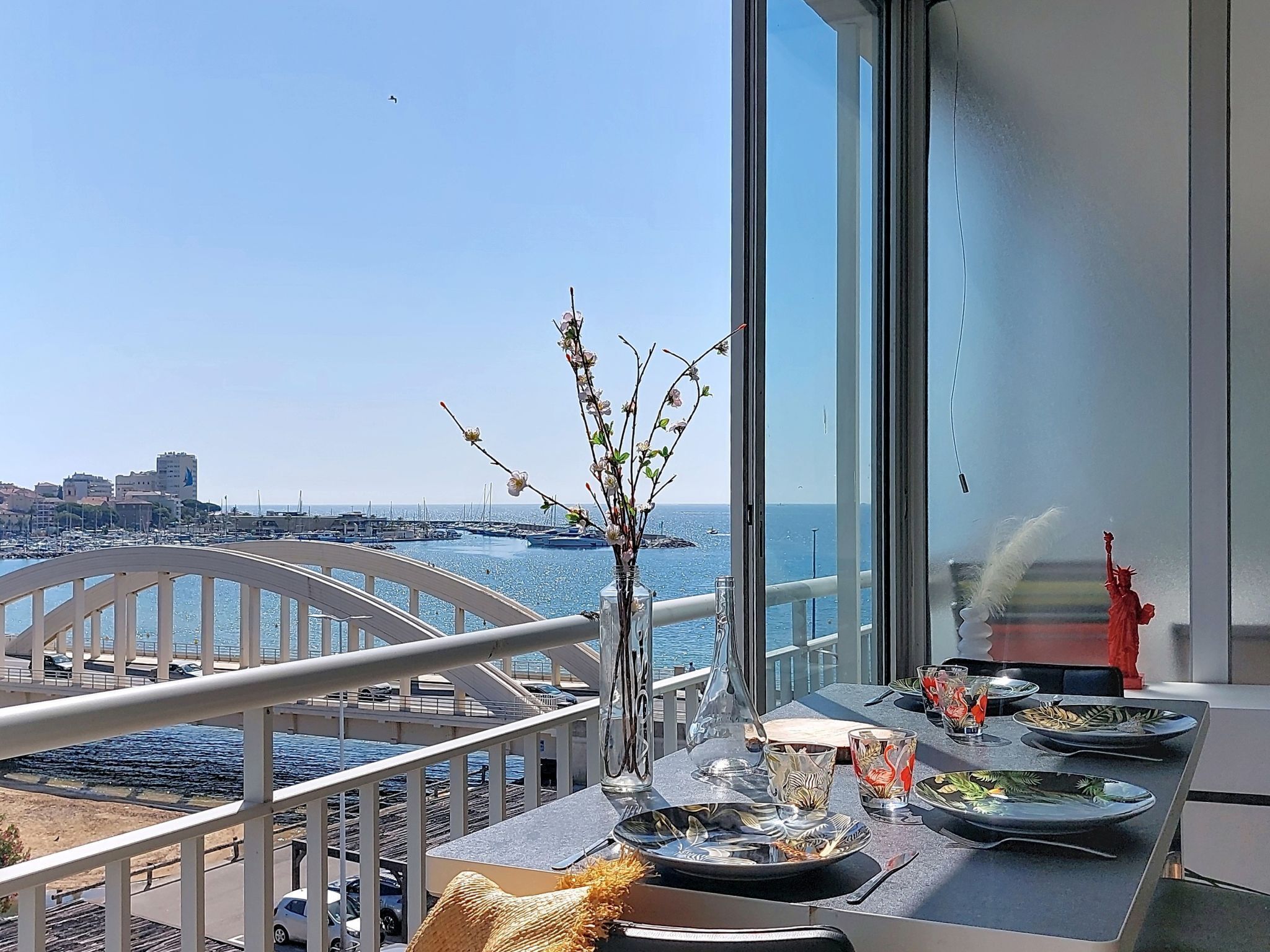 Un balcon avec vue sur la mer et la ville, un vase décoré de branches et une table mise pour le dîner.