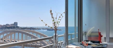 A balcony with a view of the sea and a cityscape, a vase with branches, and a table set for dining.