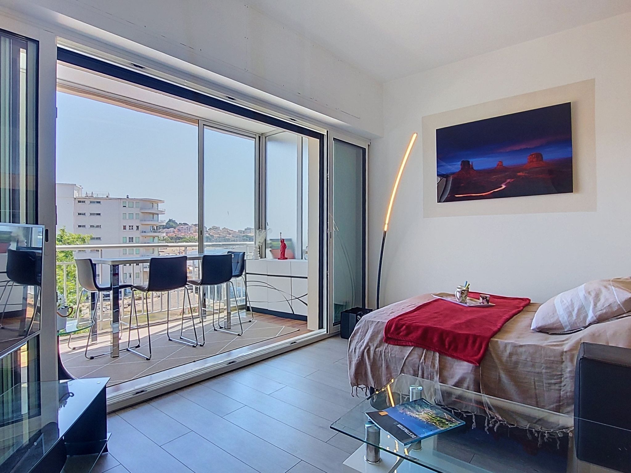 A modern room with a glass table, a bed with a red blanket, a painting, and a view of buildings through large windows.