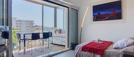 A modern room with a glass table, a bed with a red blanket, a painting, and a view of buildings through large windows.