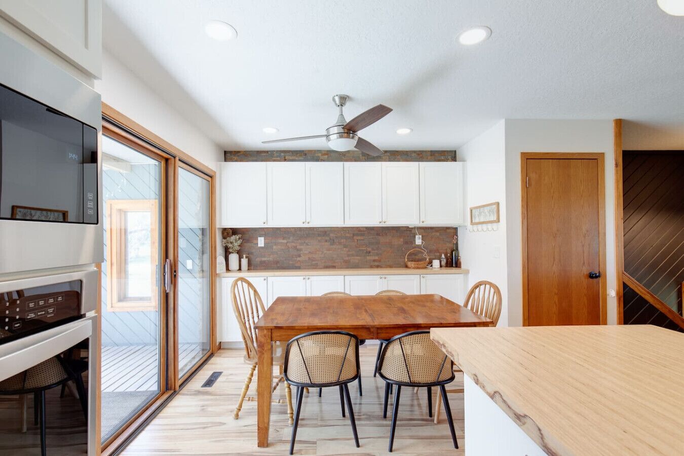 A modern kitchen with white cabinets, a wooden dining table, and a ceiling fan.