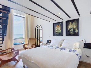 Ein Schlafzimmer mit Bett, Sessel, Nachttisch, Lampe und Meerblick.