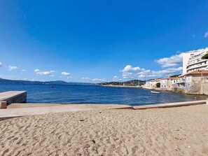 En strand med ett klart, blått hav, en sandstrand och byggnader i bakgrunden.