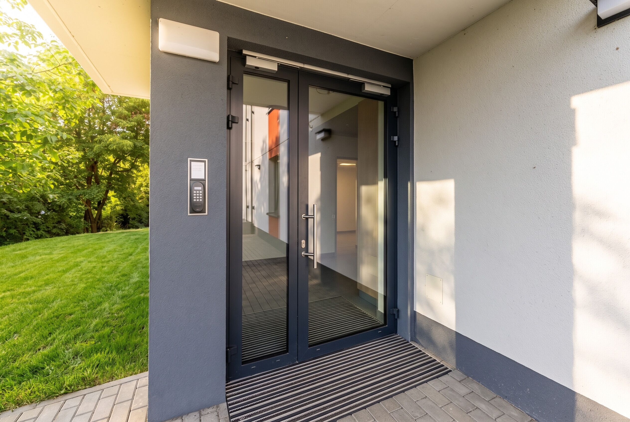 Une entrée moderne avec une porte en verre, un système de verrouillage à clavier et un petit espace extérieur agrémenté d’une pelouse.