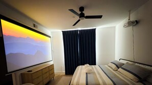 Quarto de hotel com cama, televisão, ventoinha no teto e janela com cortinas.