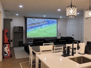 Ein Wohnzimmer mit großem Fernsehbildschirm, einer Küchenzeile, einem Esstisch und einer Sitzecke mit einer Couch.