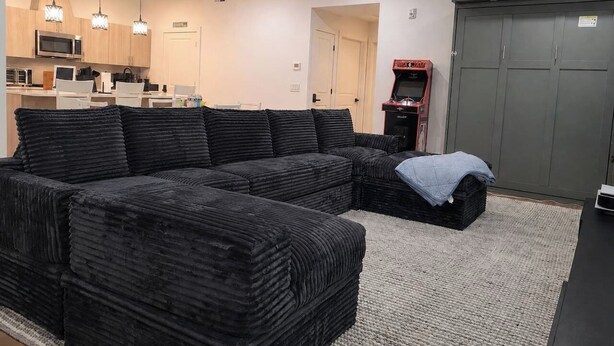 Uma sala de estar com sofá seccional, uma área de cozinha com eletrodomésticos modernos e um guarda-roupa cinza.