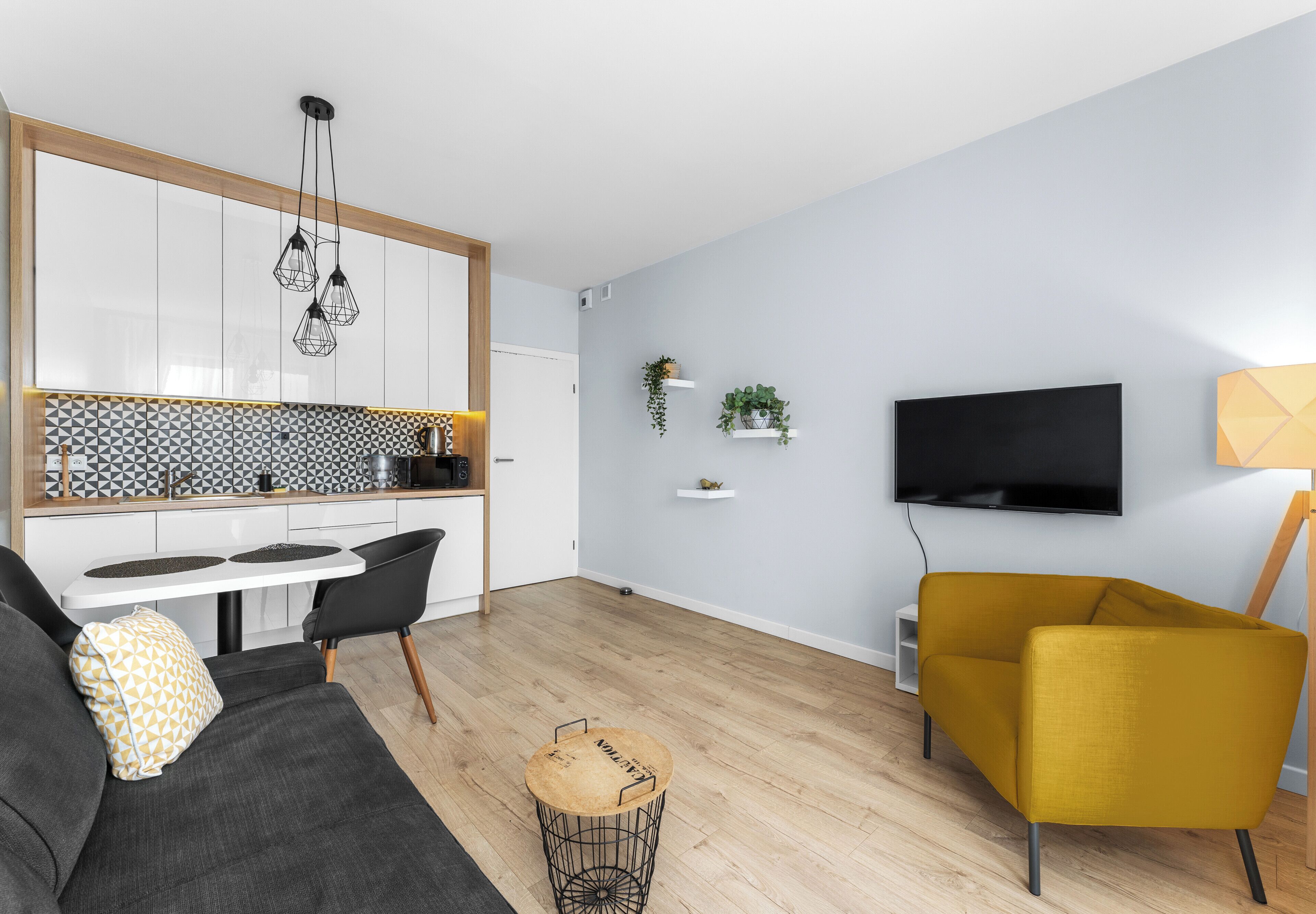 Un espai de vida modern amb un sofà, un butac armchair groc, una taula rodona petita, un televisor fixat a la paret i una zona de cuina amb un mosaique de rajoles.