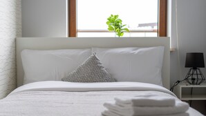 Una cama bien hecha con sábanas y almohadas blancas, una planta pequeña en el alféizar y una lámpara en la mesita de noche.
