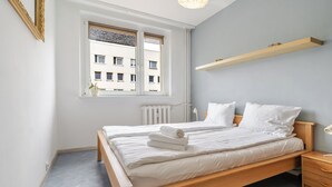 A bedroom with a wooden bed, white bedding, a window with blinds, a radiator, and a wooden shelf above the bed. - Zawodzie Homely Apartment (Katowice)