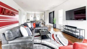 A modern living room with a sofa, coffee table, and TV. There is a kitchen in the background with a dining area. - Spacious 3bd/3ba Rooftop w/Downtown Views* Garage! (Nashville)