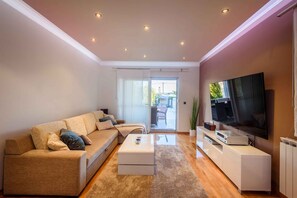 Ein modernes Wohnzimmer mit Sofa, Couchtisch, Flachbildfernseher und einer Schiebeglastür zum Außenbereich.