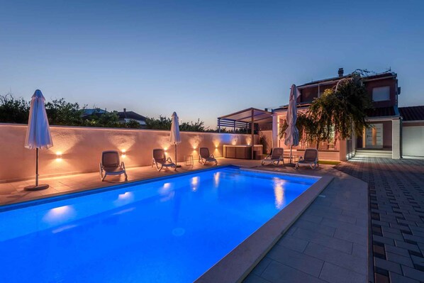A swimming pool with lounge chairs and umbrellas, a brick wall, and a building with a modern design.