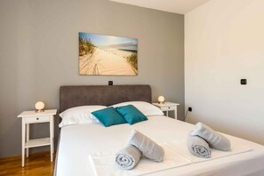 Ein ordentlich bezogenes Bett mit zwei Kissen, ein Nachttisch mit einer Lampe und ein Gemälde mit Strandmotiv an der Wand.