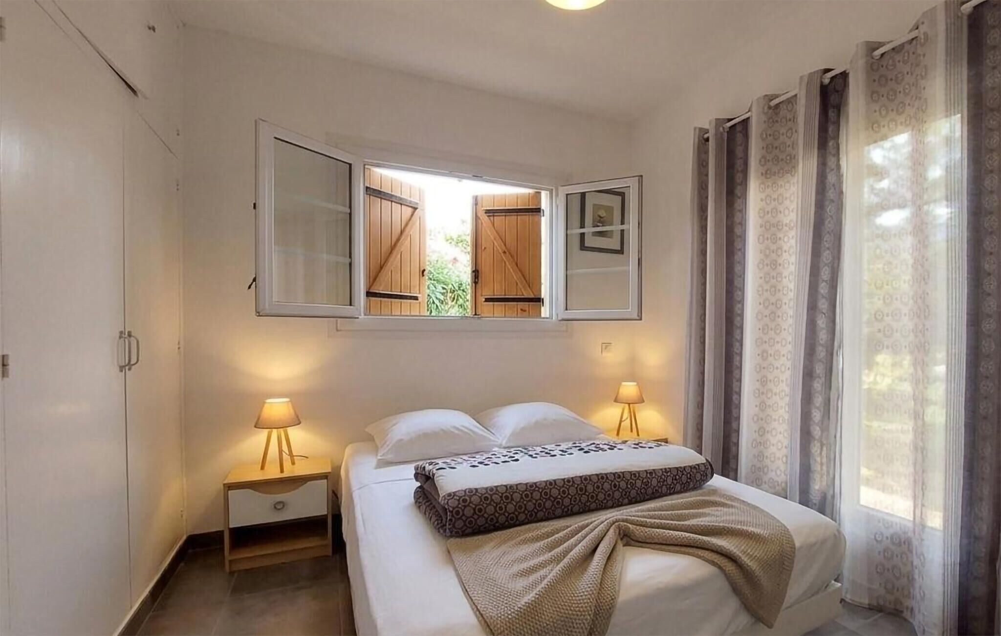 A bedroom with a bed, two bedside lamps, a window with open shutters, and a view of greenery outside.