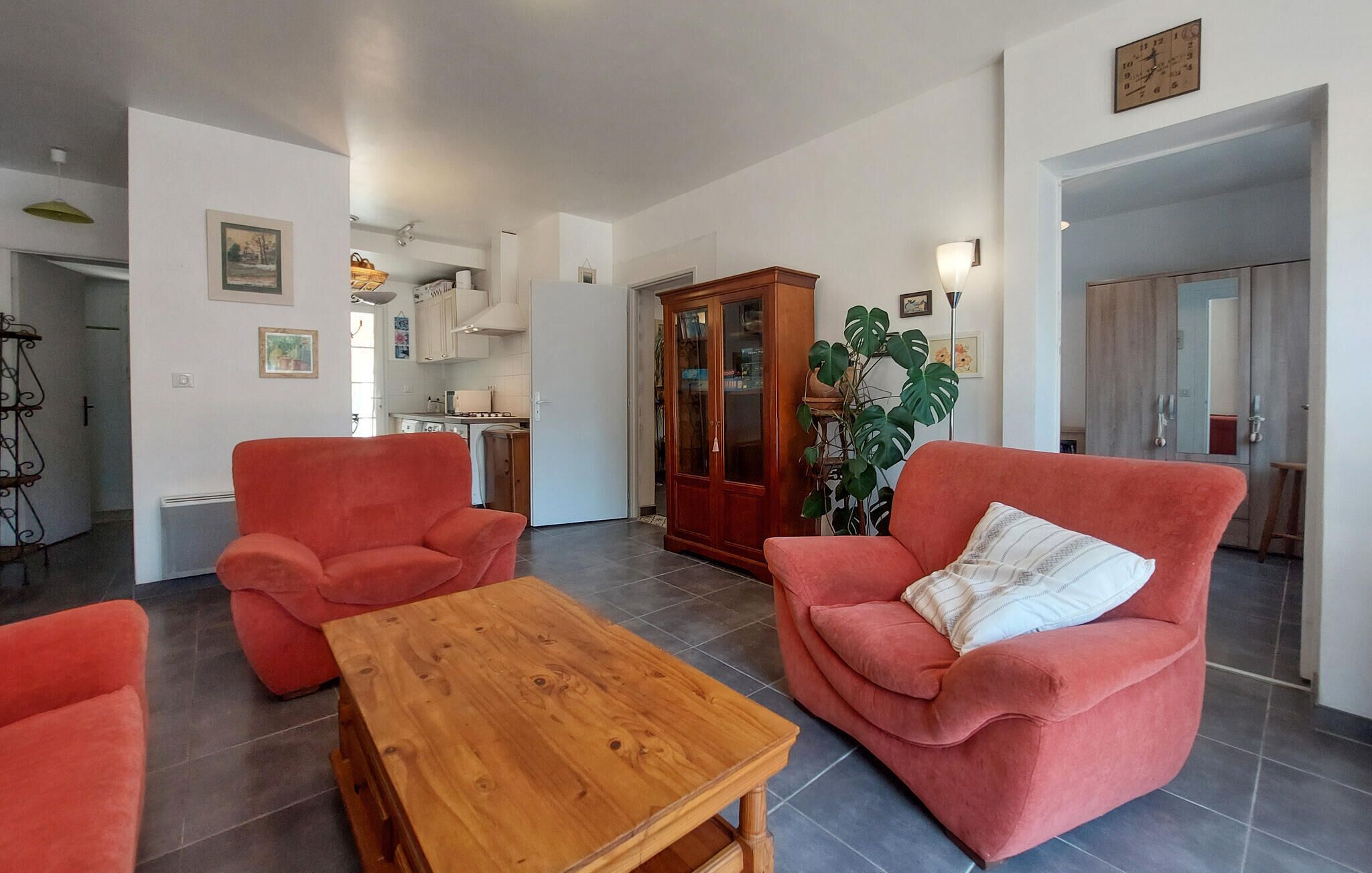 Ein Wohnzimmer mit zwei roten Sesseln, einem Holztisch, einer großen Pflanze und einer Küche im Hintergrund.