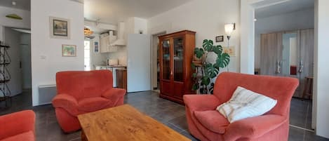Un salon avec deux fauteuils rouges, une table basse en bois, une grande plante et, au fond, une cuisine.