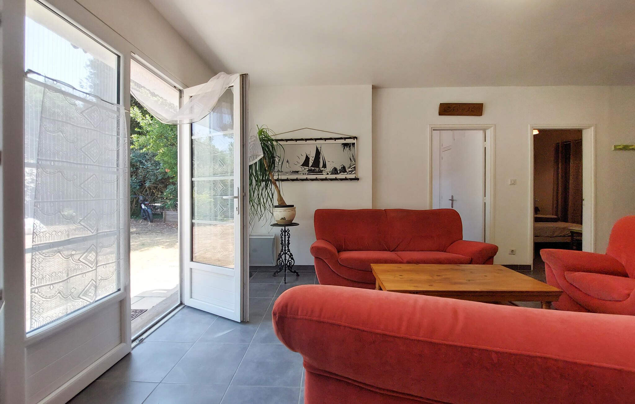 Un salon moderne doté d’une porte-fenêtre coulissante donnant sur un jardin, d’un ensemble de canapés rouges, d’une table basse en bois et d ’une œuvre d’art encadrée accrochée au mur.