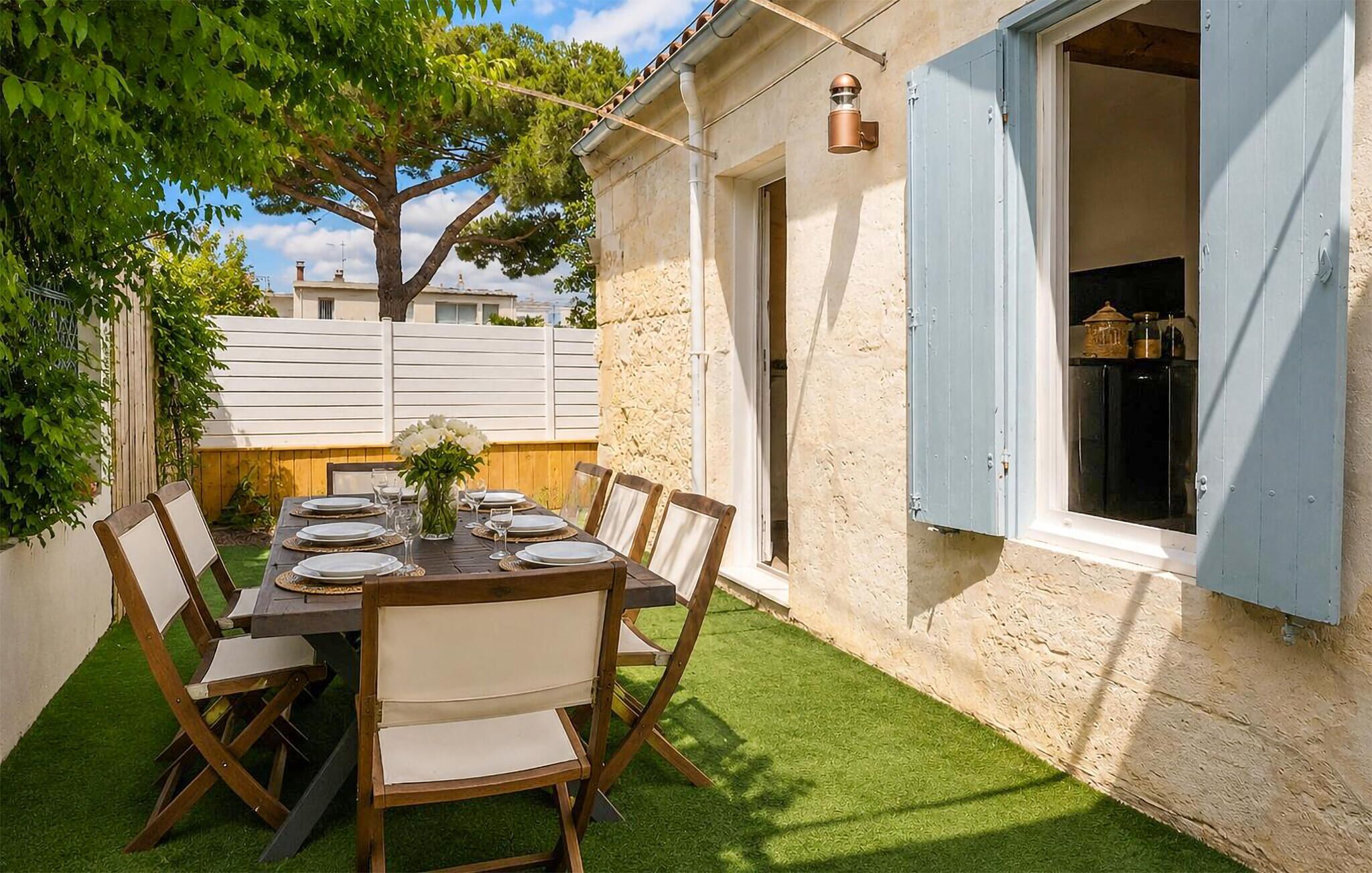 An outdoor dining area with a table set for six, surrounded by chairs, a tree, and a building with blue shutters.