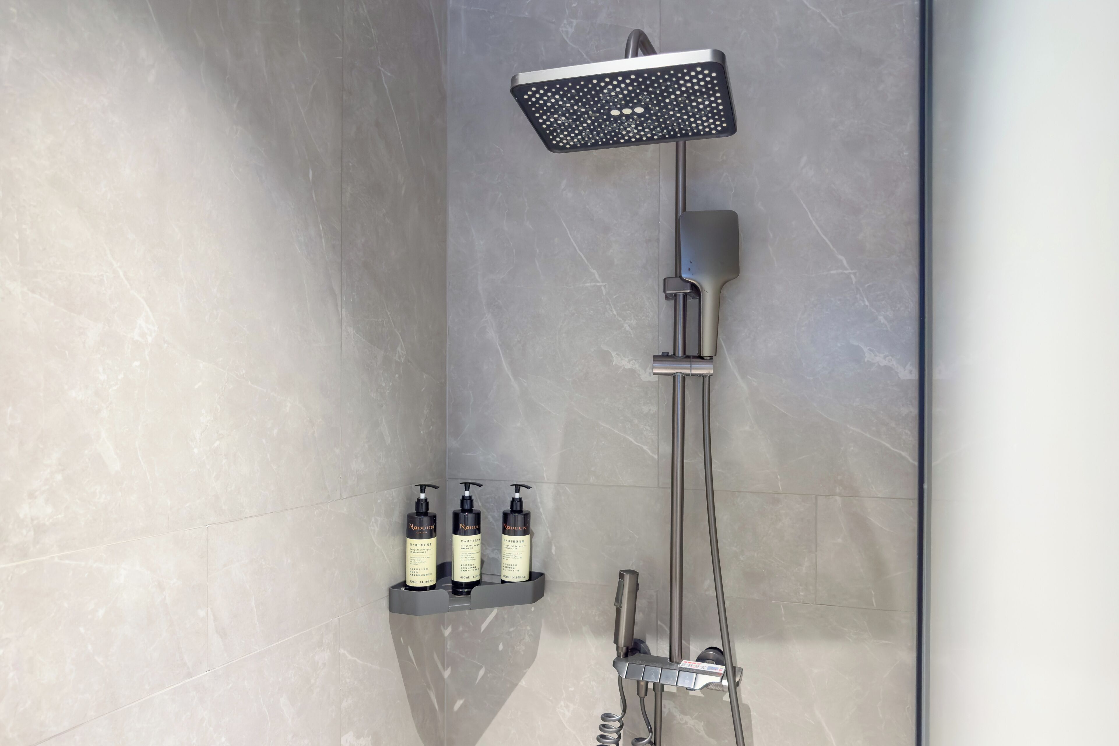 A modern shower with a handheld showerhead, a rain showerhead, and a handheld faucet. There are three bottles of shampoo on a shelf below the shower.