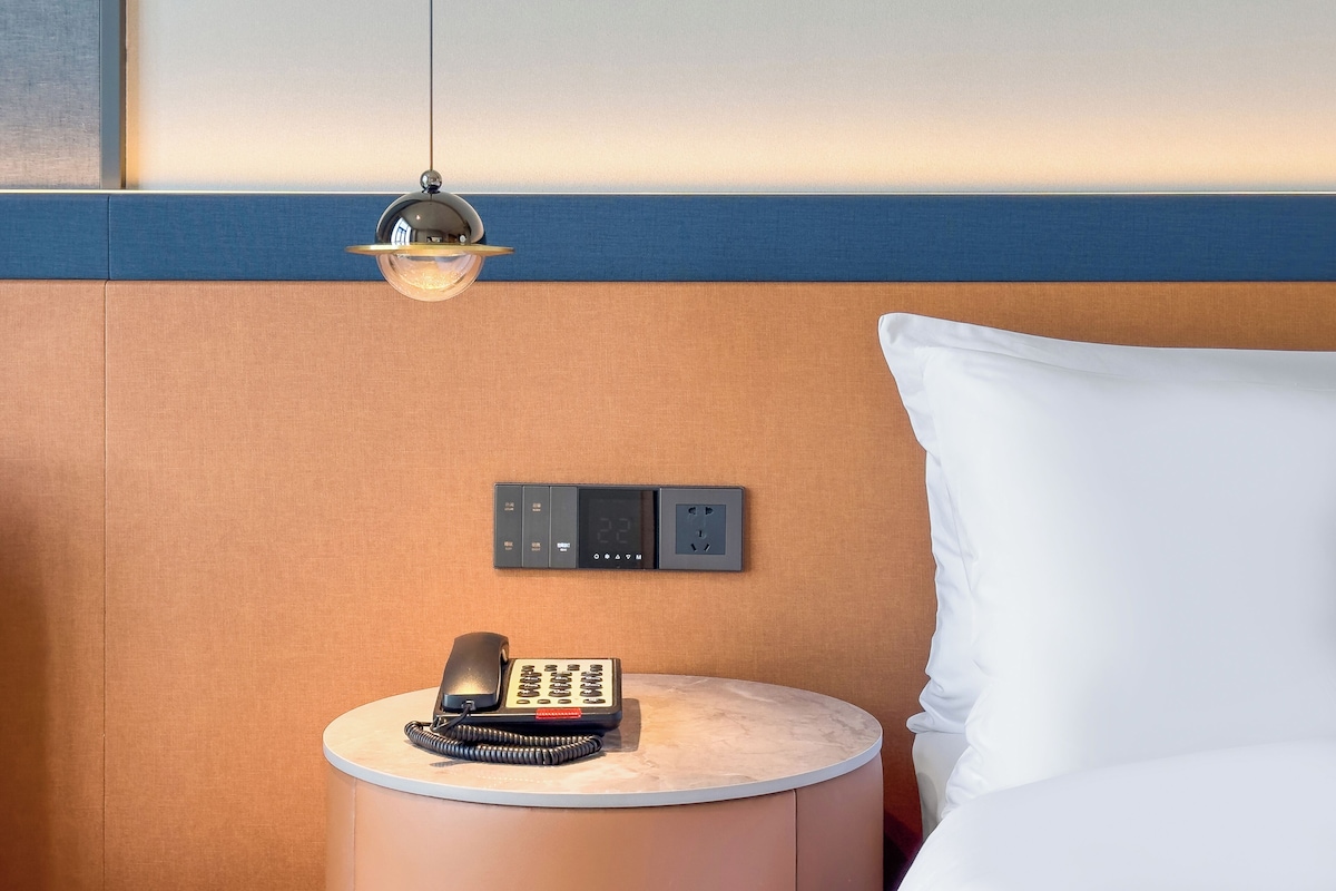 A hotel room with a bed, a bedside table with a telephone, a wall-mounted control panel, and a hanging light fixture.