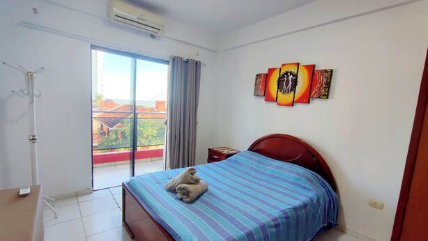 A bedroom with a bed, a bedside table, an air conditioning unit, a window with curtains, and artwork on the wall.