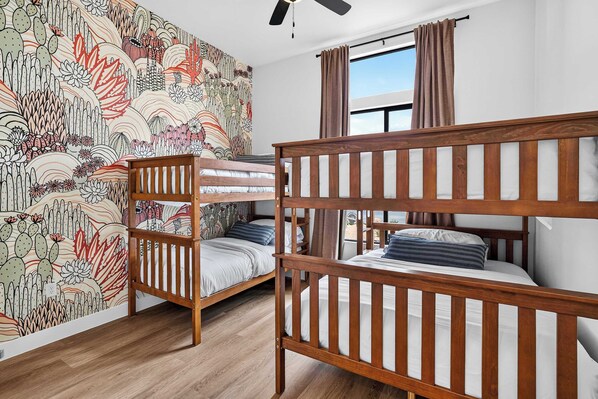 A room with bunk beds, a ceiling fan, a window with curtains, and a wall with a cactus and plant mural.