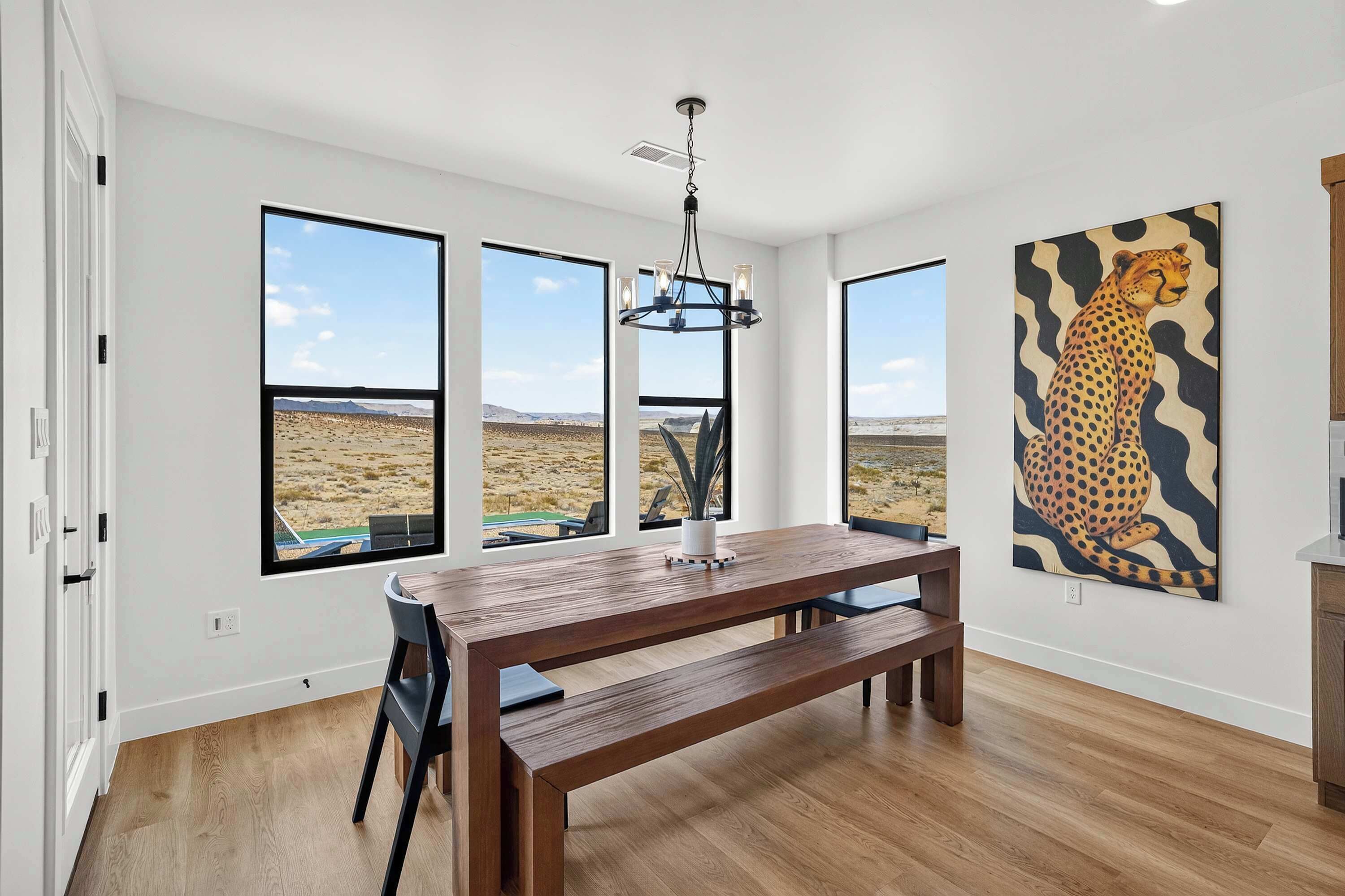Une pièce spacieuse avec une grande table à manger en bois, de nombreuses fenêtres donnant sur un paysage désertique et un tableau représentant un guépard accroché au mur.