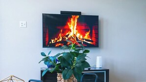 Una sala de estar con una televisión mostrando una chimenea, una mesita de madera, un sillón azul y una planta.