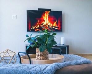 Ein Wohnzimmer mit Fernseher, der einen Kamin zeigt, einem Holztisch, einem blauen Sessel und einer Pflanze.
