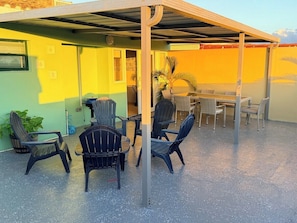 Ein Außenbereich mit Stühlen und Tischen, eine überdachte Terrasse und ein Gebäude mit grüner und gelber Fassade.