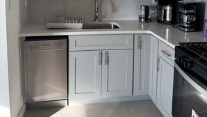 Een kleine keuken met witte kastjes, een roestvrijstalen vaatwasser, een spoelbak, een koffiezetapparaat en een fornuis.