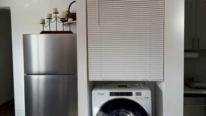 Een kleine keuken met een roestvrijstalen koelkast, een ingebouwde wasmachine en droger, en een raam met jaloezieën.