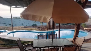 Zona de piscina al aire libre con mesa y sillas bajo un gran paraguas, rodeada por una terraza de madera y vistas al océano.