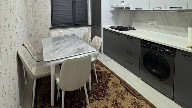 Cocina compacta con mesa de comedor, sillas, ventana con persianas, estufa empotrada, lavadora y alfombra estampada.