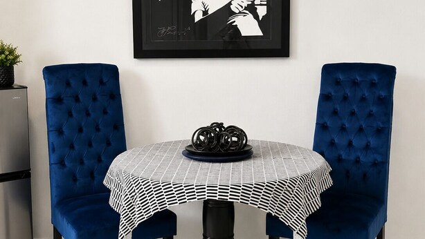Ein Esstisch mit kariertem Tischläufer, zwei blau gepolsterte Stühle und ein schwarz-weißes Bild in einem Rahmen an der Wand.