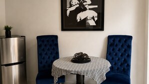 Ein Essbereich mit einem runden Tisch, zwei blau gepolsterten Stühlen, einer schwarz-weißen Tischdecke, einem gerahmten Kunstwerk darüber und einem Edelstahl-Kühlschrank zur Linken.