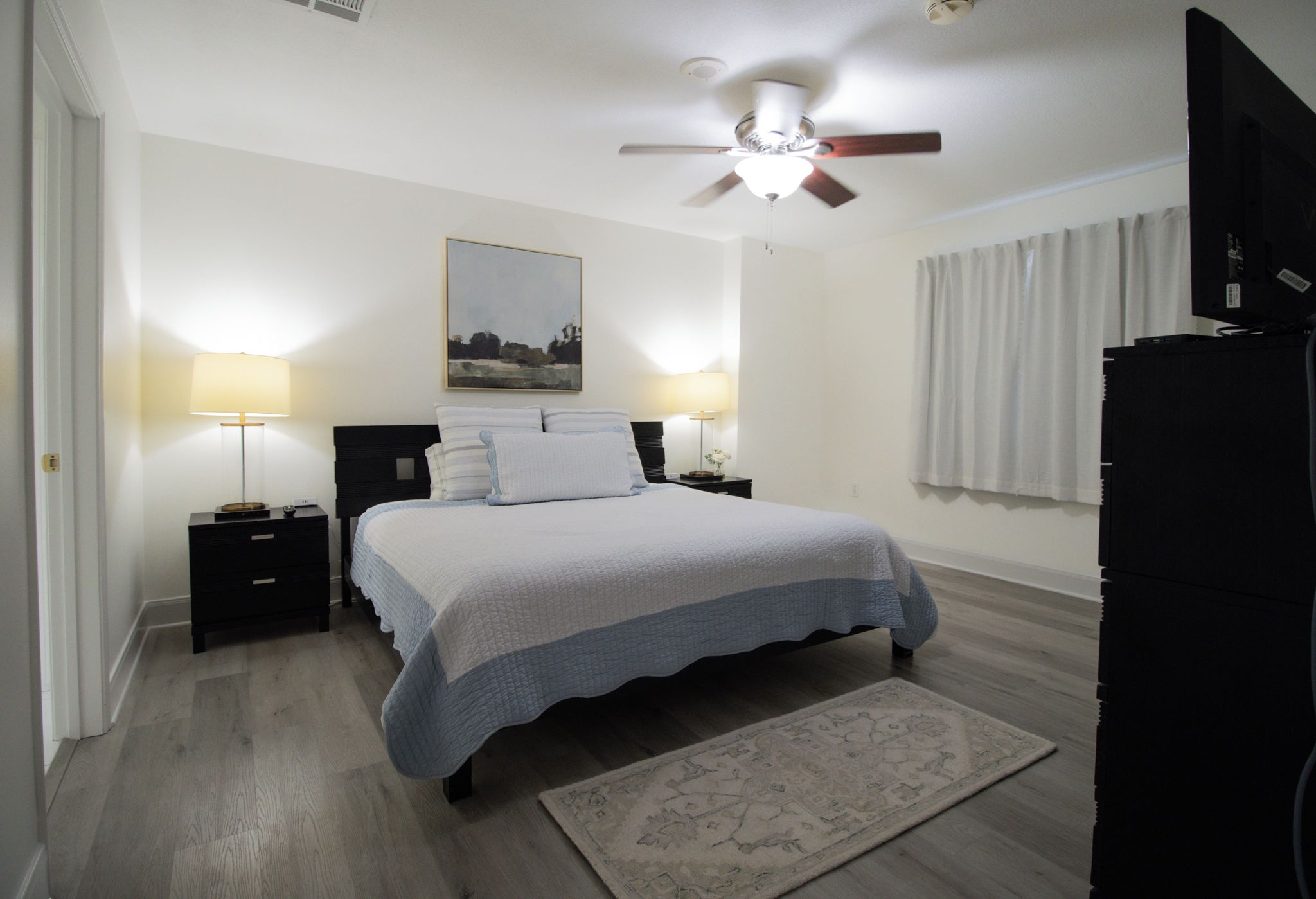 A bedroom with a bed, two bedside tables, a ceiling fan, a painting on the wall, and a television on a stand.