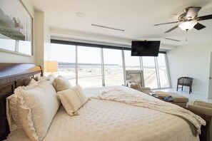 Ett hotellrum med en stor säng, en tv, ett skrivbord med en stol och utsikt över ett avlägset landskap genom stora fönster.