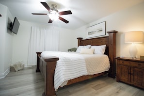 A bedroom with a wooden bed, a ceiling fan, a bedside table with a lamp, and a TV mounted on the wall.