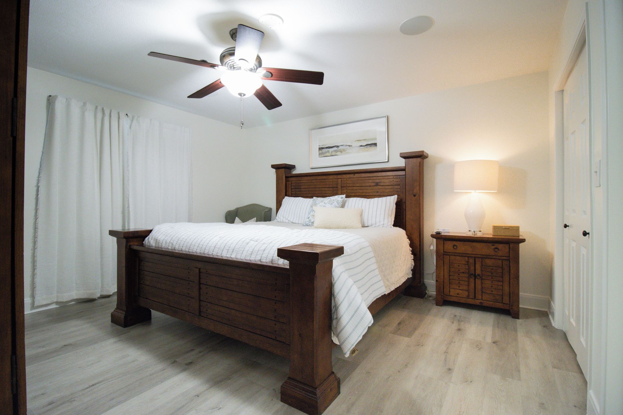 Une chambre à coucher avec un lit en bois, une table de chevet avec une lampe, un ventilateur de plafond et un tableau encadré au mur.