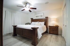 A bedroom with a wooden bed, a nightstand with a lamp, a ceiling fan, and a framed picture on the wall.