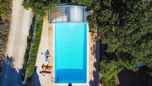Une vue aérienne d’une piscine rectangulaire entourée de verdure, avec un chemin pavé et une structure couverte à proximité.
