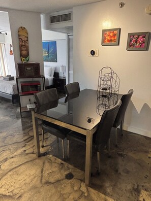 A dining area with a glass-top table, four chairs, a microwave, a small cabinet, and a decorative item on the wall. - Chic studio condo with Free Parking & AC in vibrant Downtown Honolulu (Honolulu)