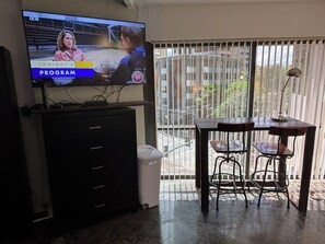 A television mounted on a wall displaying a program, a desk with chairs, and a window with blinds.