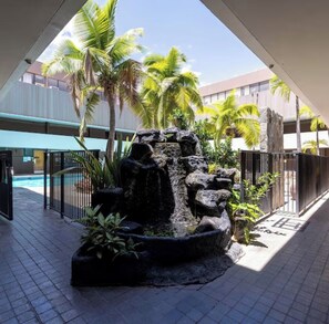 An indoor garden with a rock feature, palm trees, and a pool in the background.