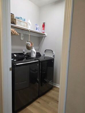 A compact laundry room with a washing machine, dryer, and shelves with cleaning supplies. - Merchants Quarter 4BR Pet Friendly Beach Gear Propane BBQ (Galveston)